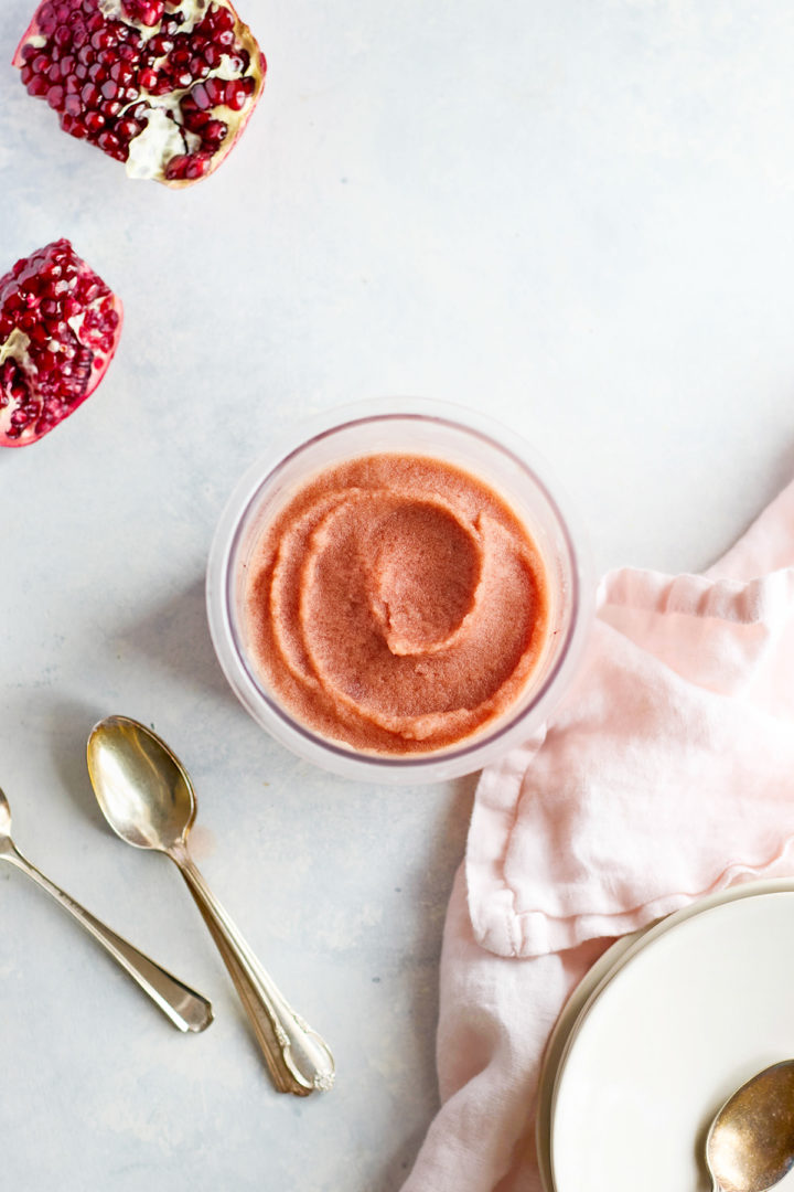Overhead view of smooth, pink pomegranate sorbet swirled in a freezer-safe container. A halved pomegranate and two silver spoons rest nearby on a light background.