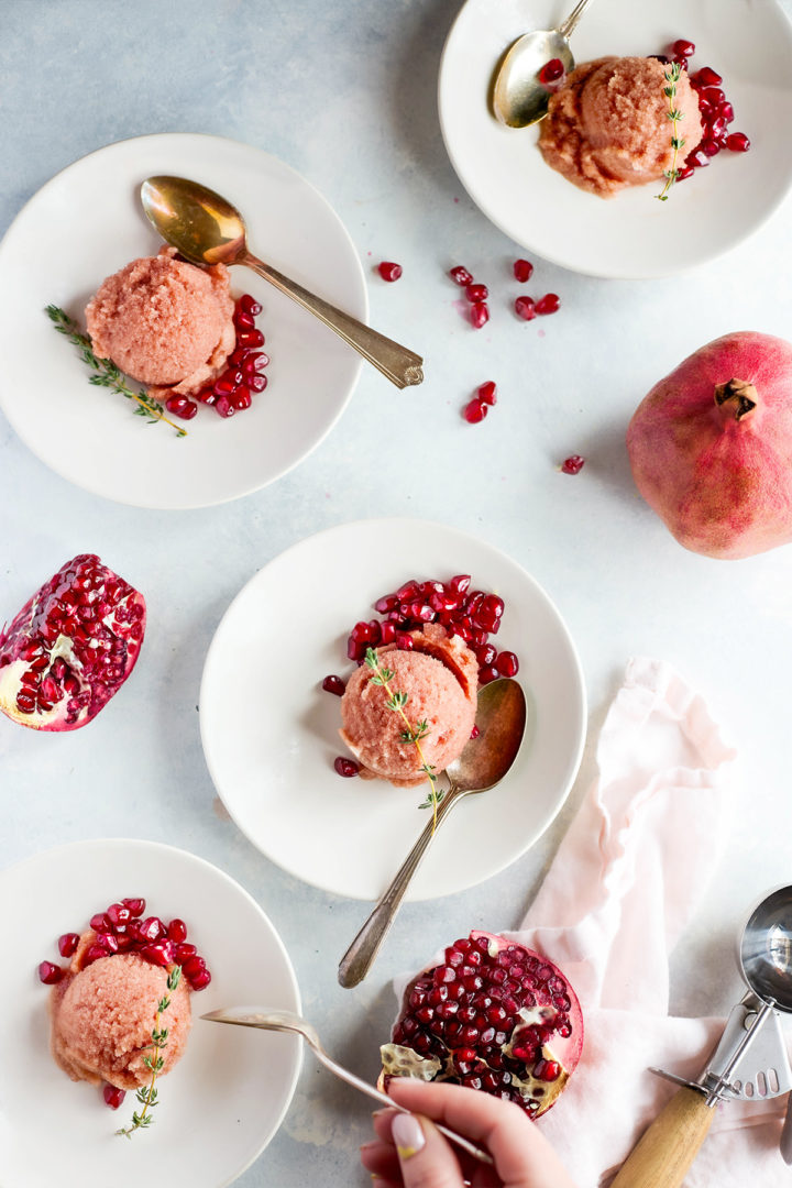 Four white plates each hold a scoop of homemade pomegranate sorbet, garnished with pomegranate seeds and fresh thyme. A hand reaches toward one plate.

