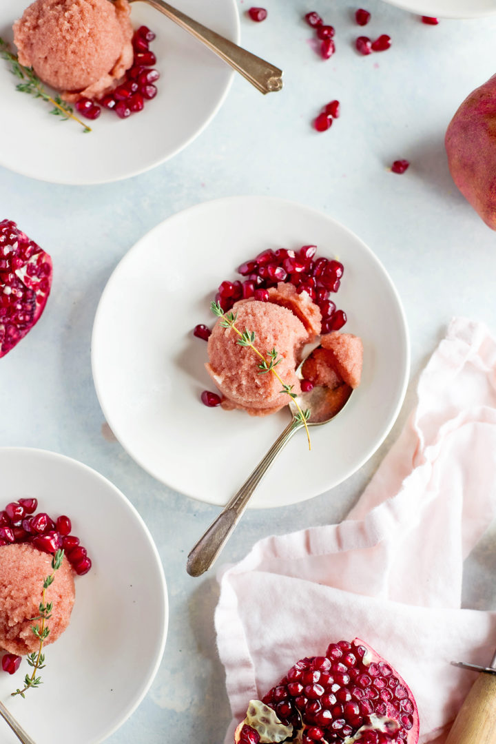 A silver spoon digs into a soft scoop of easy pomegranate sorbet, served with pomegranate arils and a sprig of thyme on a white plate.