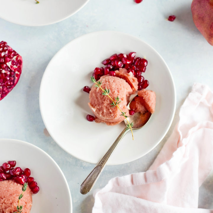 Four white dessert plates arranged on a light surface, each with a scoop of pink pomegranate sorbet garnished with pomegranate seeds and a sprig of fresh thyme. A halved pomegranate, soft pink napkin, and metal scoop frame the scene, creating a vibrant and inviting display of a refreshing pomegranate dessert.