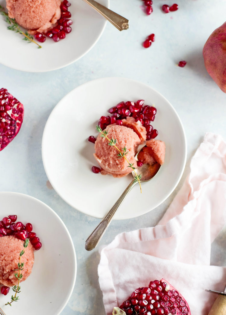 Four white dessert plates arranged on a light surface, each with a scoop of pink pomegranate sorbet garnished with pomegranate seeds and a sprig of fresh thyme. A halved pomegranate, soft pink napkin, and metal scoop frame the scene, creating a vibrant and inviting display of a refreshing pomegranate dessert.