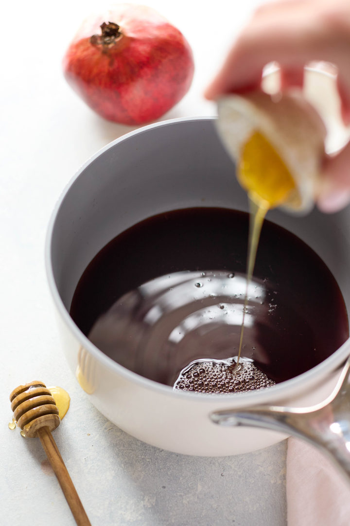 Honey is being drizzled into a saucepan filled with deep red pomegranate juice. A whole pomegranate and honey dipper sit nearby on a pale surface.