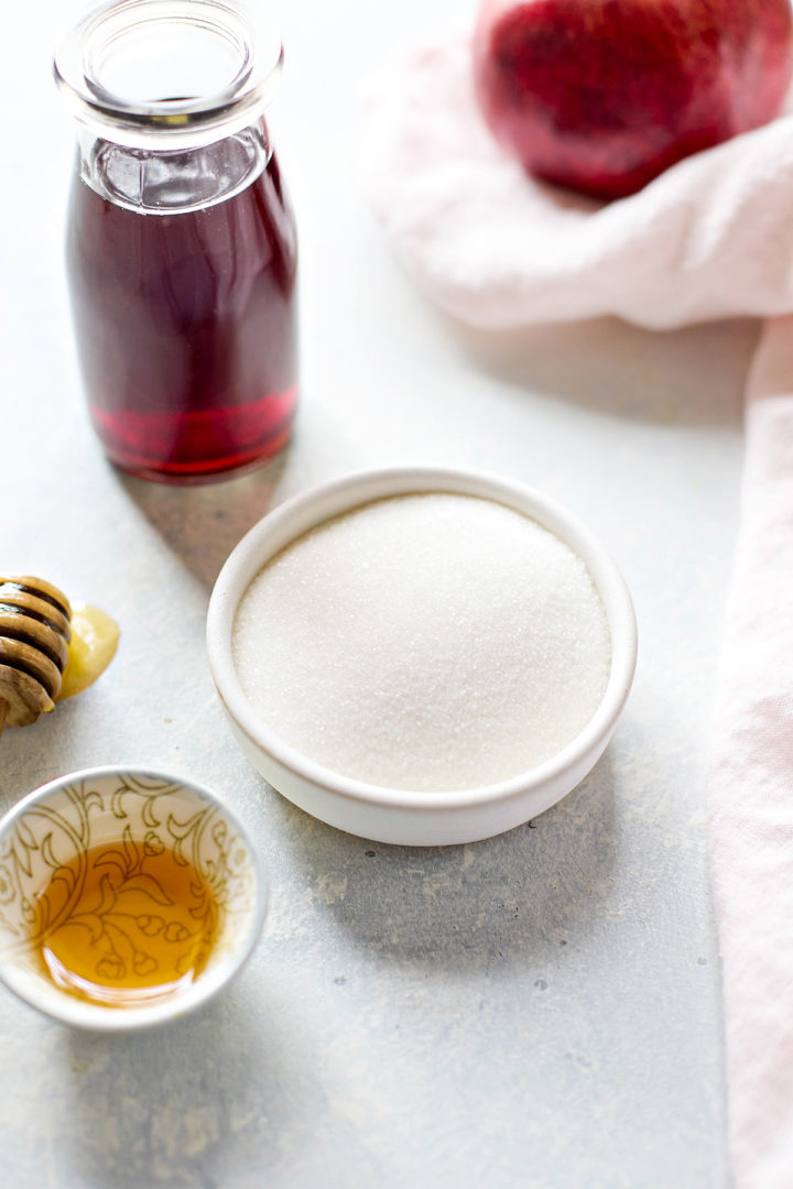 Ingredients for pomegranate sorbet arranged on a counter: a bowl of granulated sugar, a small dish of honey, and a bottle of dark red pomegranate juice.