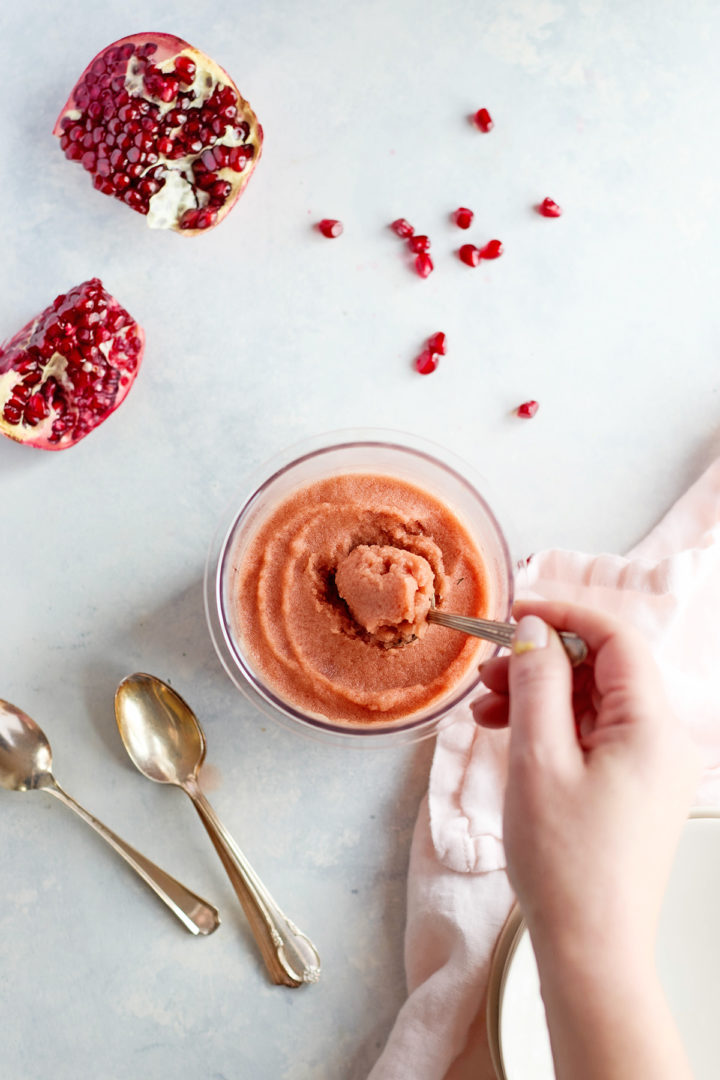 A hand holding a spoon scoops pomegranate sorbet from a chilled container. Juicy pomegranate seeds are scattered above, next to halved fruit and vintage silver spoons.