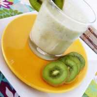 A pineapple orange smoothie in a glass, on an orange plate with kiwi slices.