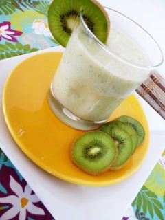 A pineapple orange smoothie in a glass, on an orange plate with kiwi slices.