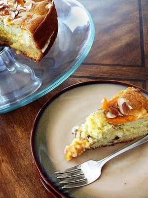 A slice of apricot almond cake on a dessert plate, next to a cake stand with the rest of the cake. 