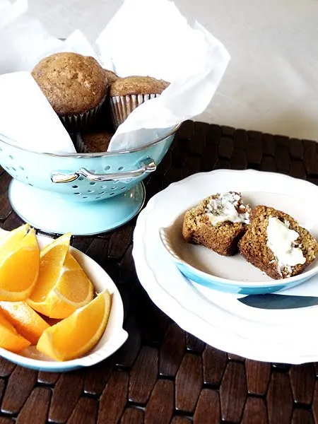 A colander piled with ginger carrot muffins, a dish of orange slices, and a muffin cut in half and slathered with butter.