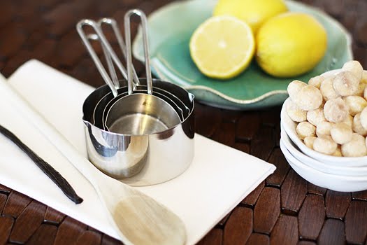 Measuring cups, halved lemons, and macadamia nuts on a dark surface to use in this white chocolate blondie recipe. 