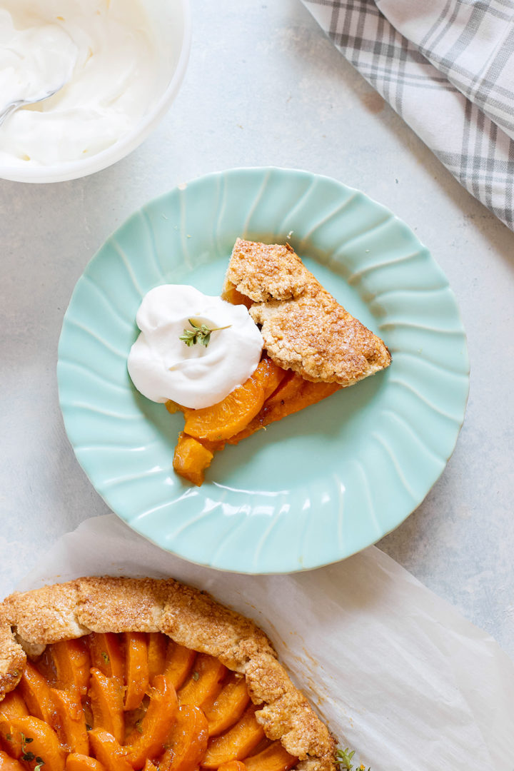 Close-up of a single slice of apricot galette on a mint-green plate, topped with whipped cream and a thyme sprig. Part of the full galette and a bowl of whipped cream are visible in the background.