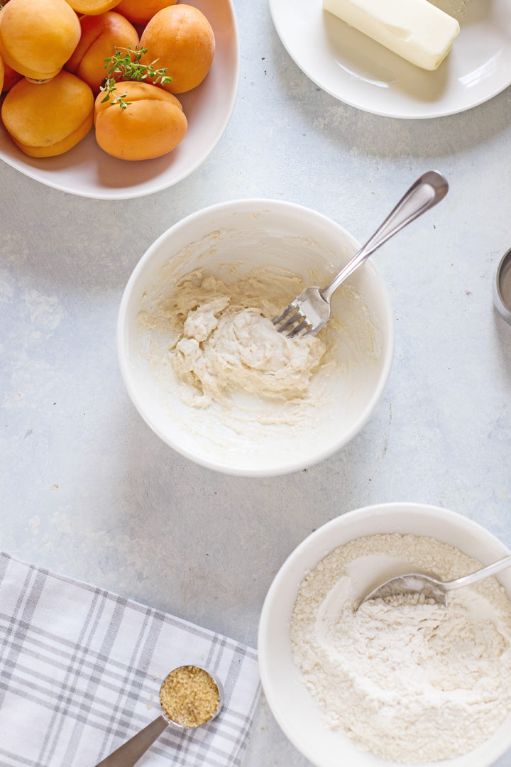 A mixing bowl of ingredients sits next to a bowl of flour, whole apricots, and a plate with butter. A spoonful of coarse sugar rests on a cloth napkin.