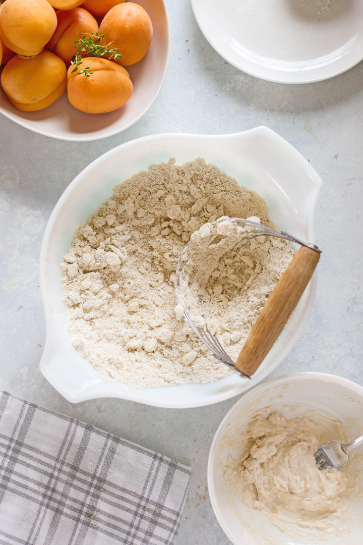 Overhead view of a bowl of flour and butter mixture being cut with a pastry blender. Nearby, another bowl holds a creamy dough mixture, and a third dish contains whole apricots with sprigs of fresh thyme.