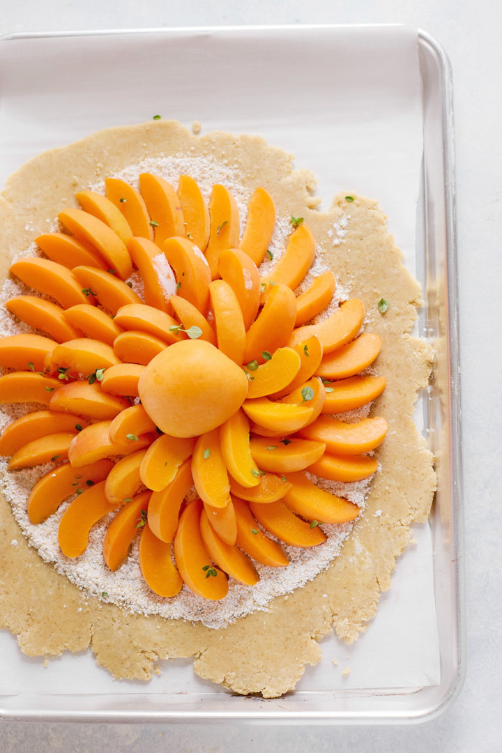 Unbaked apricot galette on a baking sheet. The crust is rolled out in a rustic circle, and fresh apricot slices are artfully layered in a flower shape, surrounded by almond meal.

