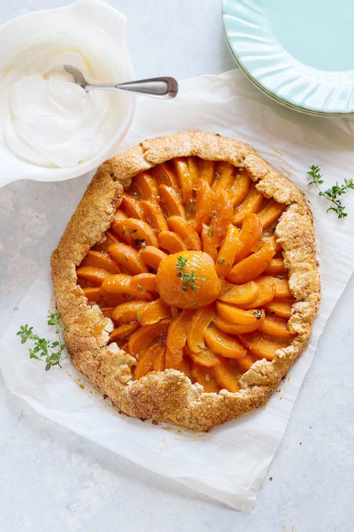 Baked apricot galette with golden crust and a glossy, spiral arrangement of apricot slices, centered around a halved apricot with thyme garnish. A dish of whipped cream sits nearby.