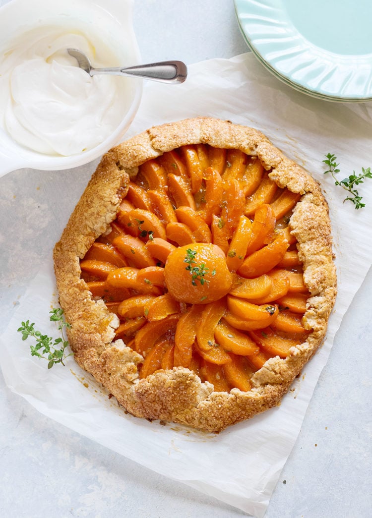 Baked apricot galette with golden crust and a glossy, spiral arrangement of apricot slices, centered around a halved apricot with thyme garnish. A dish of whipped cream sits nearby.