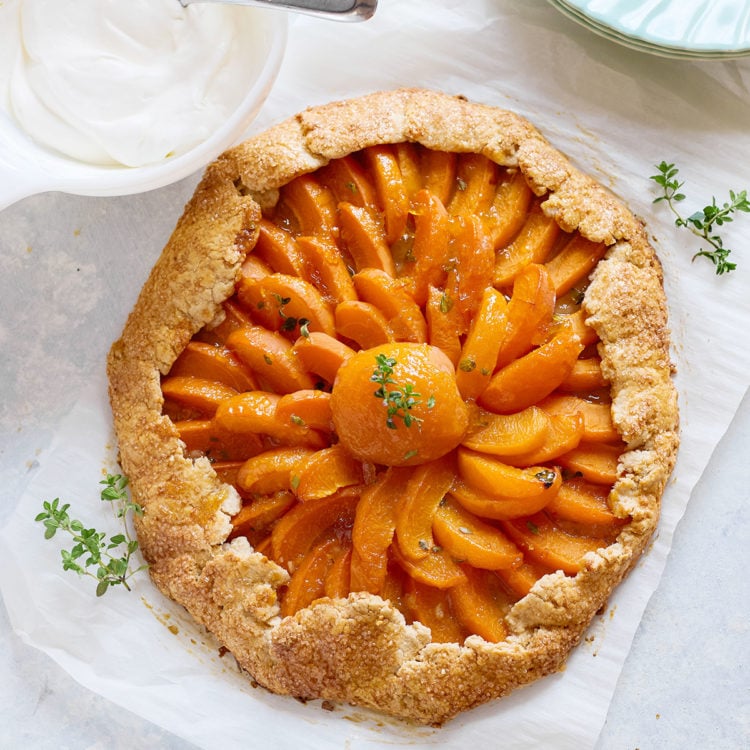 Baked apricot galette with golden crust and a glossy, spiral arrangement of apricot slices, centered around a halved apricot with thyme garnish. A dish of whipped cream sits nearby.