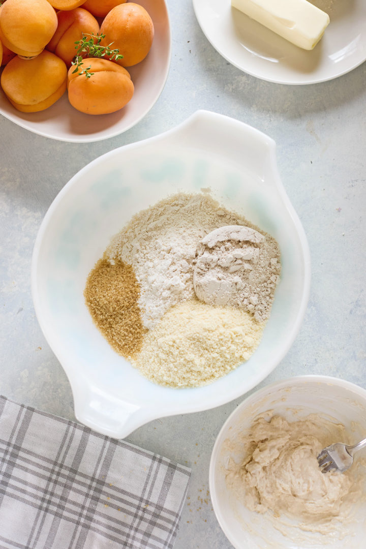 Dry ingredients for crust preparation, including flour, almond meal, and coarse sugar, arranged in a white mixing bowl. A second bowl contains a wet dough mixture.