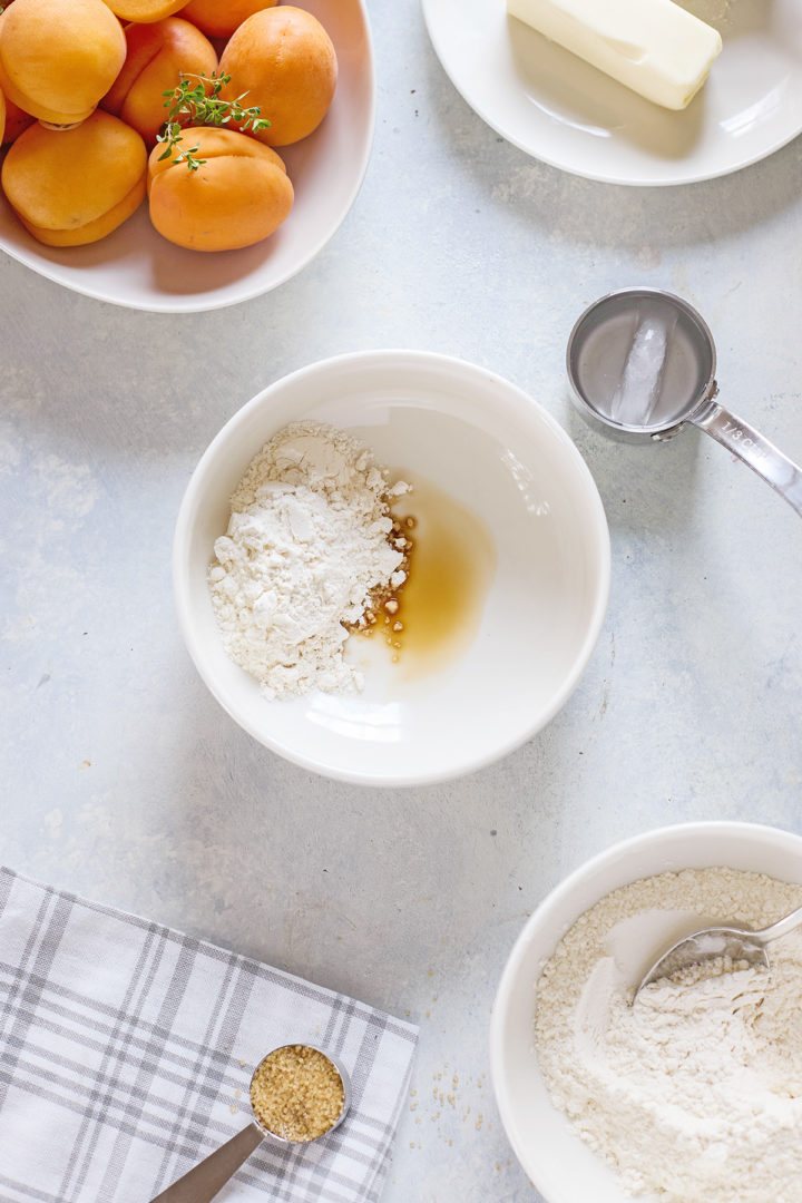 A bowl of flour with a splash of vanilla and a plate of butter nearby. Coarse sugar and more flour rest on a plaid cloth, alongside a bowl of apricots with sprigs of thyme.