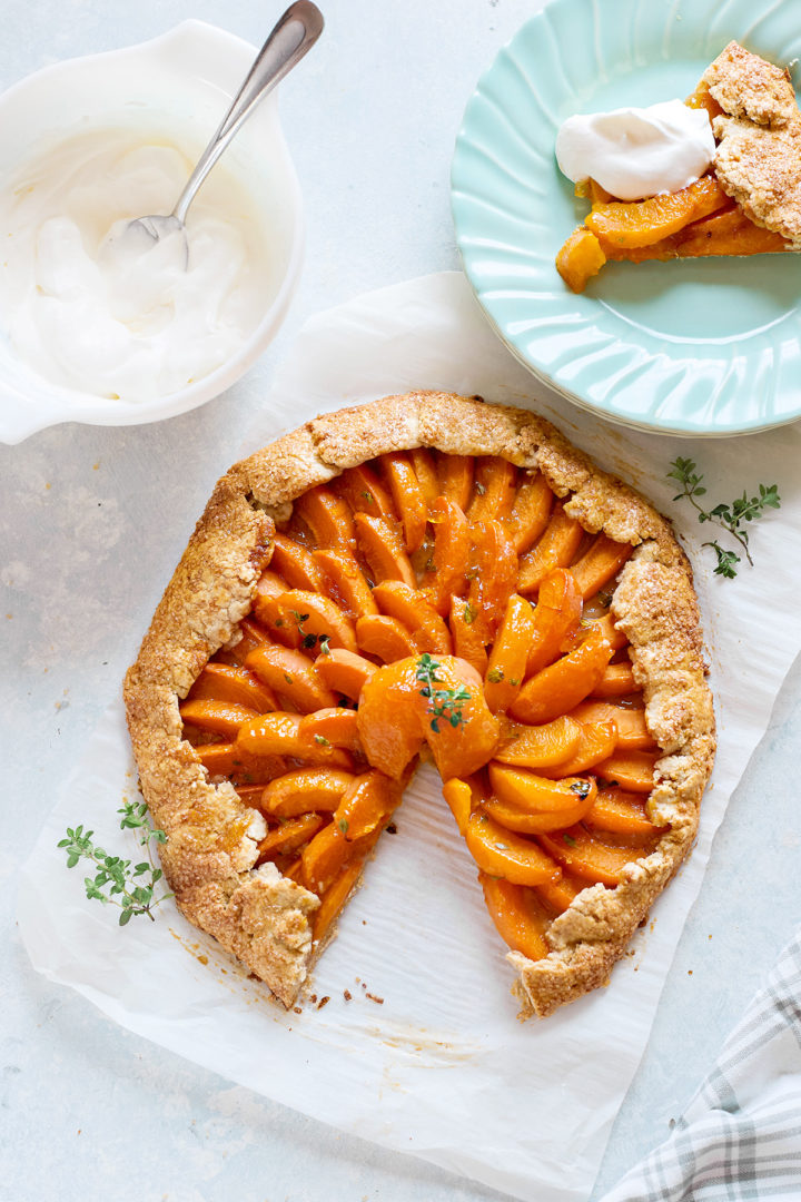 Partially sliced apricot galette on parchment paper. One slice is served on a mint-green plate with a dollop of whipped cream. The galette’s crust is crisp and sugared.