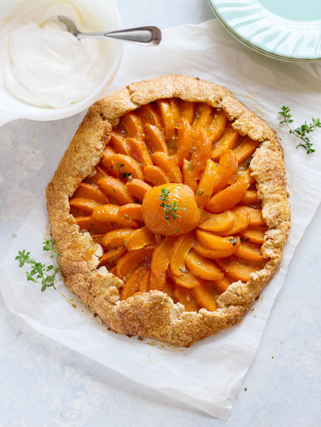 Baked apricot galette with golden crust and a glossy, spiral arrangement of apricot slices, centered around a halved apricot with thyme garnish. A dish of whipped cream sits nearby.