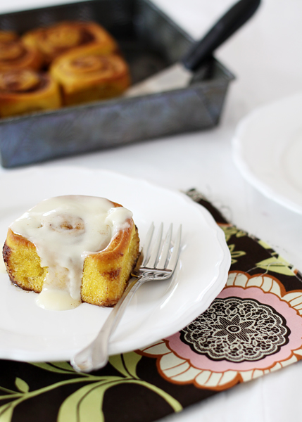 photo of a plate with a pumpkin cinnamon roll with cream cheese frosting next to a pan of freshly baked pumpkin rolls
