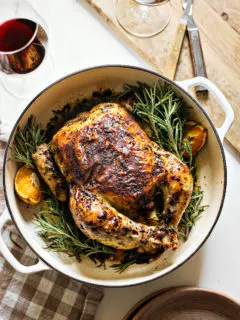 photo of rosemary roasted chicken in a white cast iron roasting pan with fresh rosemary and oranges net to dinner plates, glasses of red wine, and a cutting board