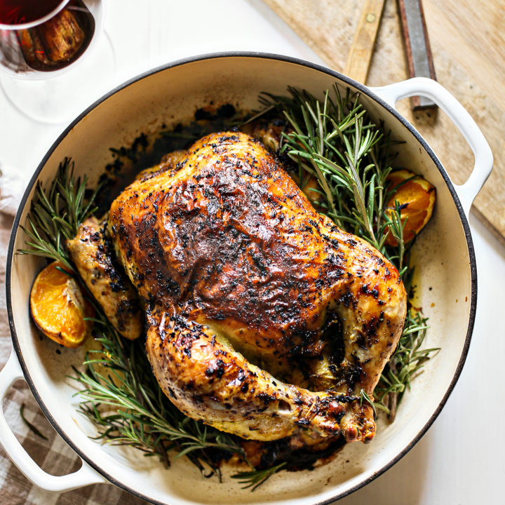 photo of rosemary roasted chicken in a white cast iron roasting pan with fresh rosemary and oranges net to dinner plates, glasses of red wine, and a cutting board