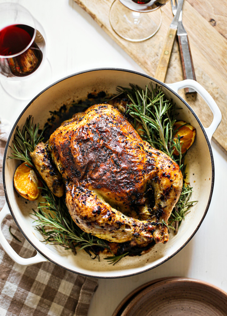 photo of rosemary roasted chicken in a white cast iron roasting pan with fresh rosemary and oranges net to dinner plates, glasses of red wine, and a cutting board