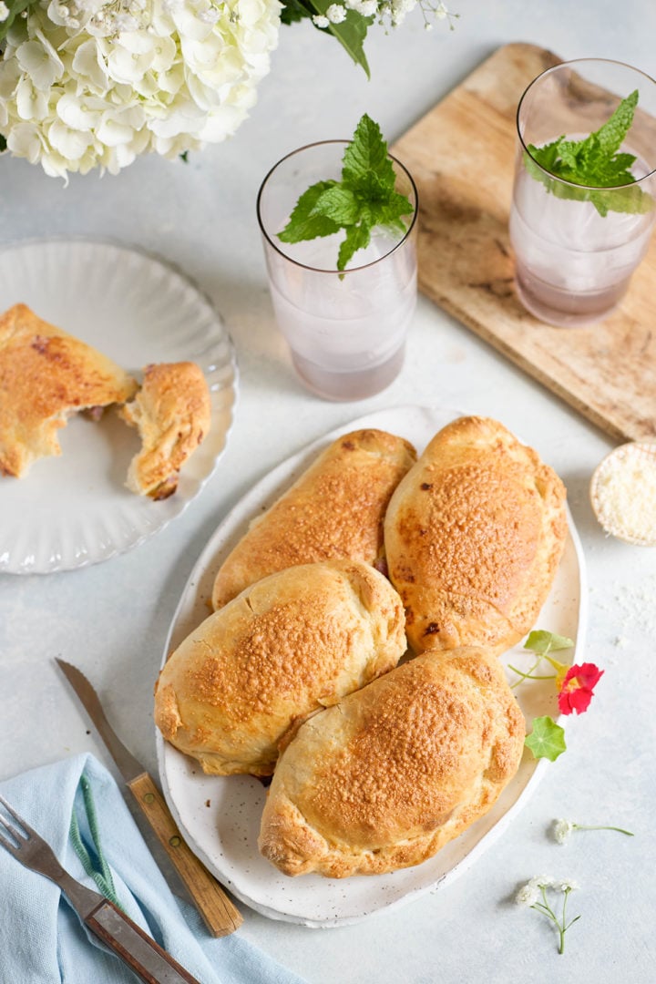 Overhead view of a platter with five baked ham and cheese calzones. Two iced drinks with mint sit nearby, along with a fork, knife, and pale blue cloth napkin.
