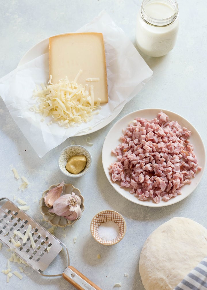 Flat lay of ham and cheese calzone ingredients: shredded cheese, diced ham, mustard, garlic cloves, coarse salt, and pizza dough on a light counter with a striped napkin.