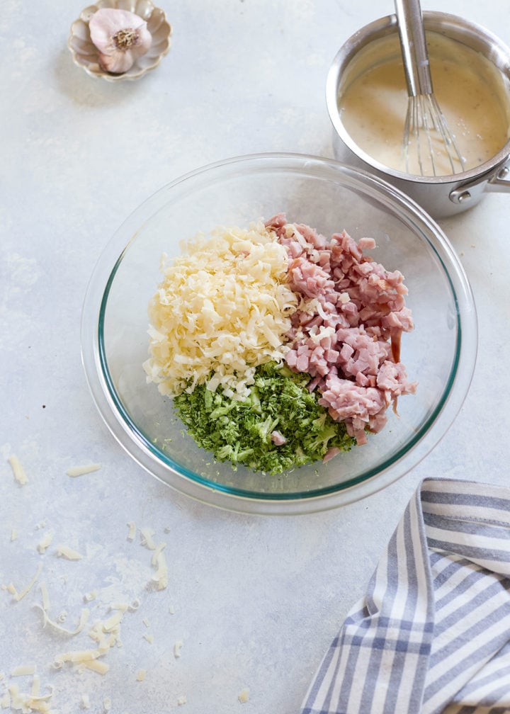 Glass mixing bowl with piles of shredded cheese, chopped ham, and finely chopped broccoli before mixing. A saucepan of creamy sauce sits in the background on the counter.