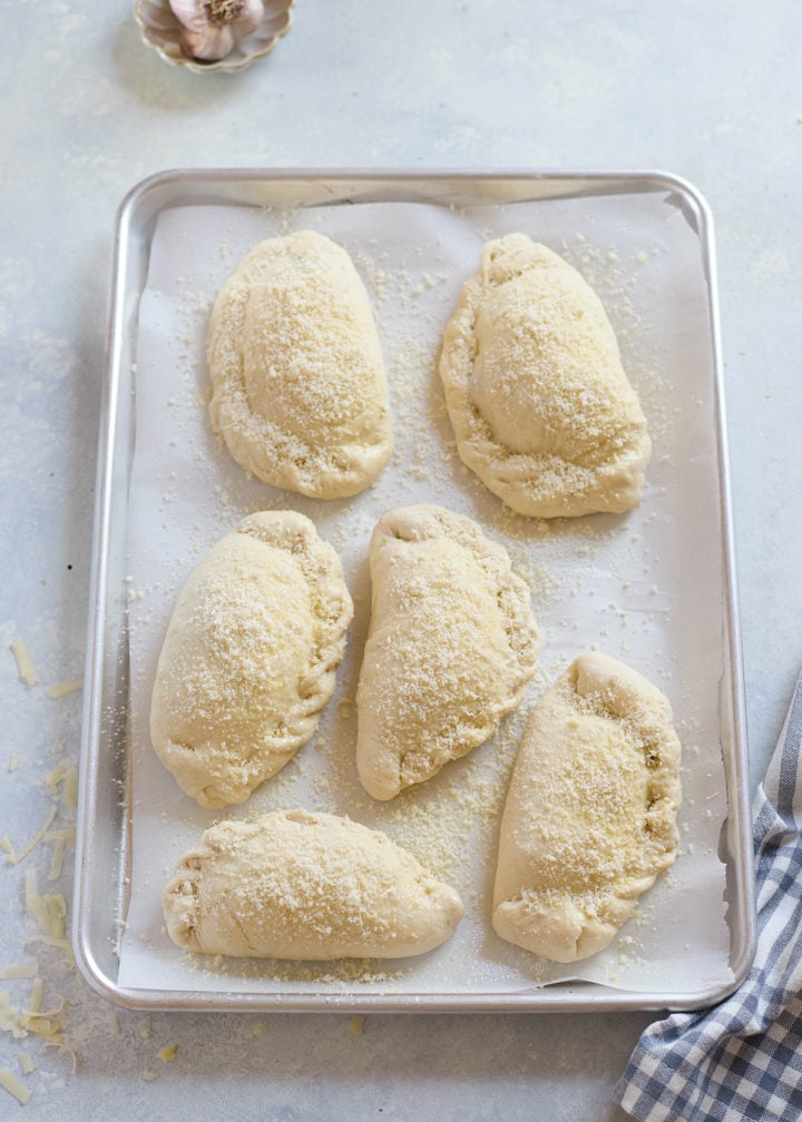 Six unbaked ham and cheese calzones arranged on a parchment-lined baking sheet. Each calzone is folded and sprinkled with shredded cheese, ready to bake.