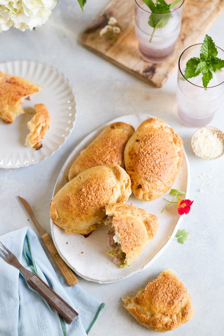 Table scene with a platter of golden brown baked ham and cheese calzones. One calzone is cut open to reveal filling, surrounded by iced drinks with mint and fresh flowers.