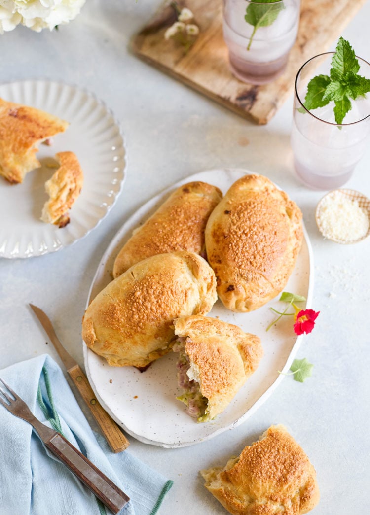 Table scene with a platter of golden brown baked ham and cheese calzones. One calzone is cut open to reveal filling, surrounded by iced drinks with mint and fresh flowers.