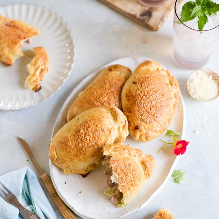 Table scene with a platter of golden brown baked ham and cheese calzones. One calzone is cut open to reveal filling, surrounded by iced drinks with mint and fresh flowers.