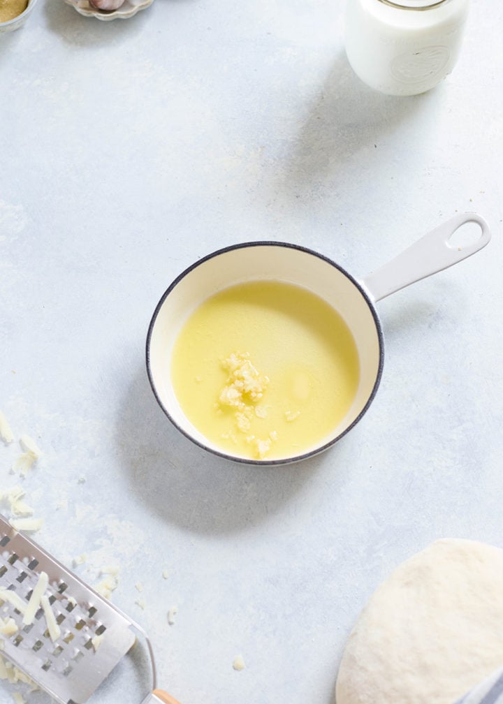 Small saucepan filled with melted butter and minced garlic on a light countertop. A batch of dough and shredded cheese are nearby, with a garlic bulb in a dish in the top corner.