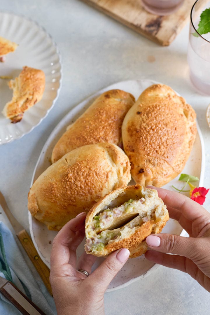 Close-up of hands holding a baked ham and cheese calzone broken open to show the creamy ham and broccoli filling. Three whole calzones rest on an oval plate in the background.