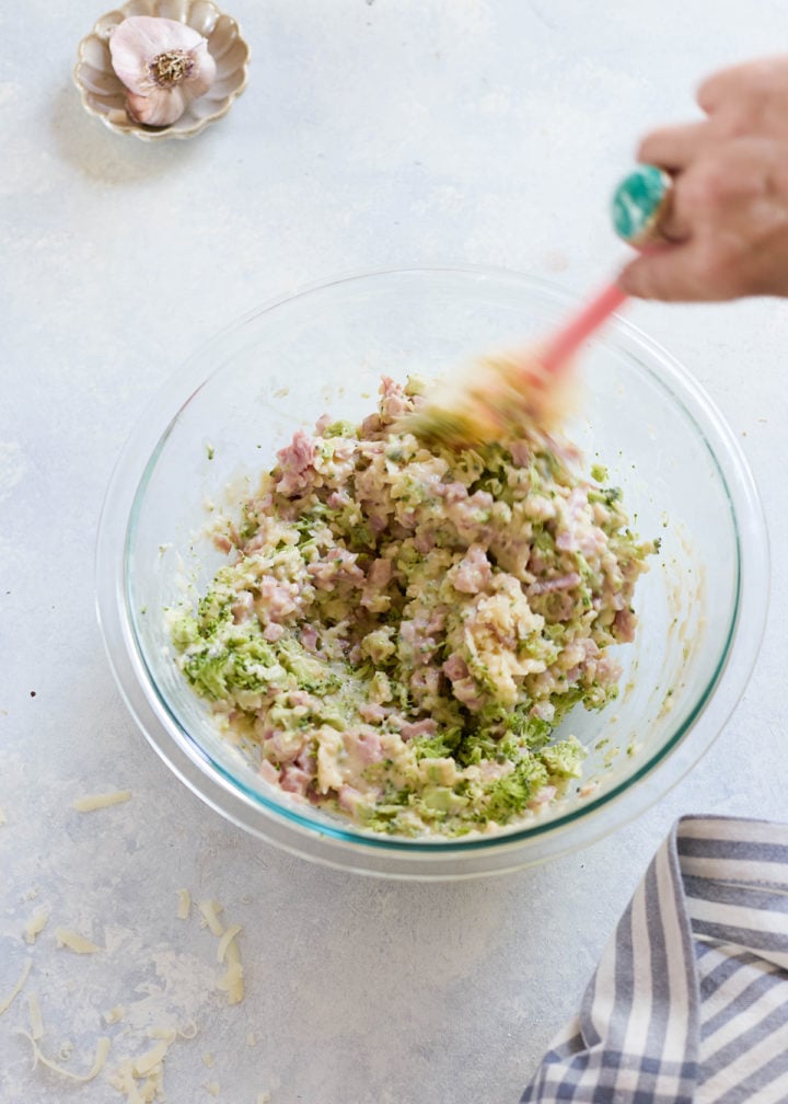A hand stirs a glass bowl filled with chopped ham, shredded cheese, and finely chopped broccoli, combining the calzone filling with a pink spatula. A garlic bulb sits in a small dish nearby.