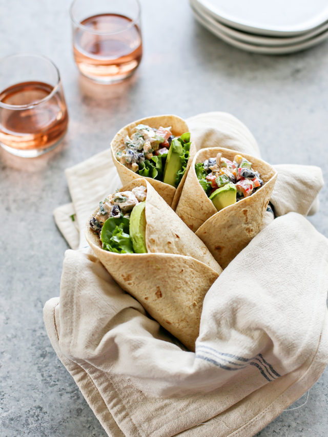 A linen-lined basket holds three Southwest Chicken Salad Wraps with creamy chicken salad, lettuce, and avocado. Two glasses of rosé and white plates sit in the background on a gray surface.