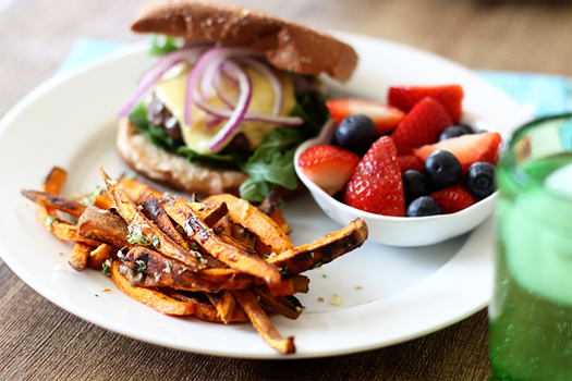 a white plate with fruit salad, baked sweet potato fries, and a burger