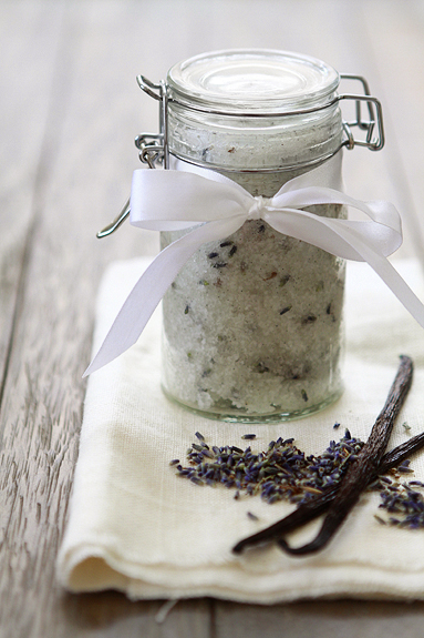 A small glass jar of vanilla sugar scrub. A vanilla bean and lavender rest in the foreground. 