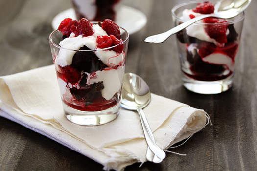 Two raspberry brownie trifle in glasses with spoons on a dark wooden table