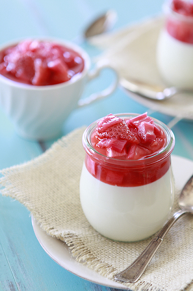 A glass jar of vanilla bean panna cotta topped with strawberry rhubarb compote. More compote is in a tea cup in the background. 