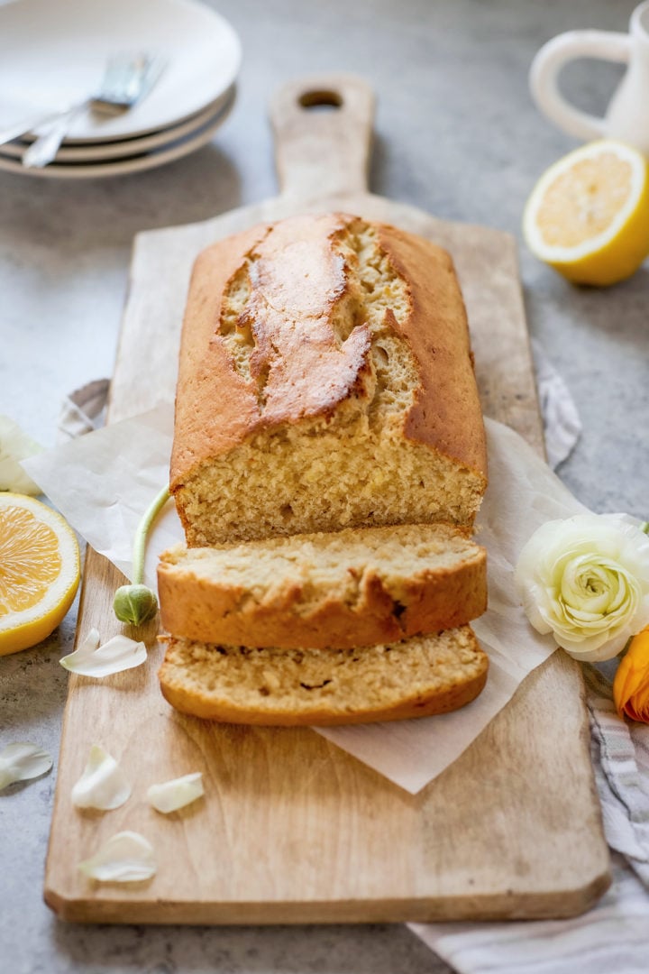 A close-up of a sliced lemon banana bread loaf showing its moist interior texture. Set on a wooden board with lemon halves and white and yellow flowers surrounding it—this homemade banana bread with lemon looks soft and fluffy.