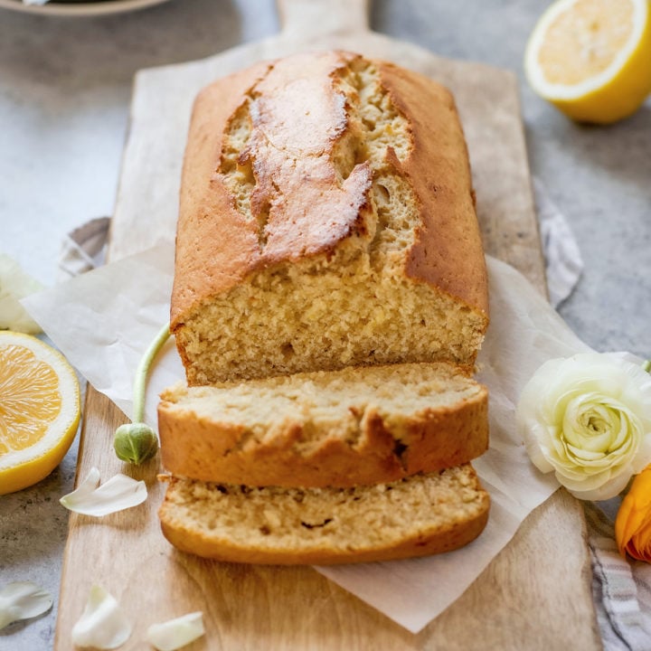 A loaf of banana bread with lemon juice sits on a wooden cutting board, sliced to reveal its soft, moist crumb. Surrounded by fresh lemon halves, white flower petals, and a light springtime setting, this lemon banana bread looks bright and inviting—perfect for gifting or brunch.