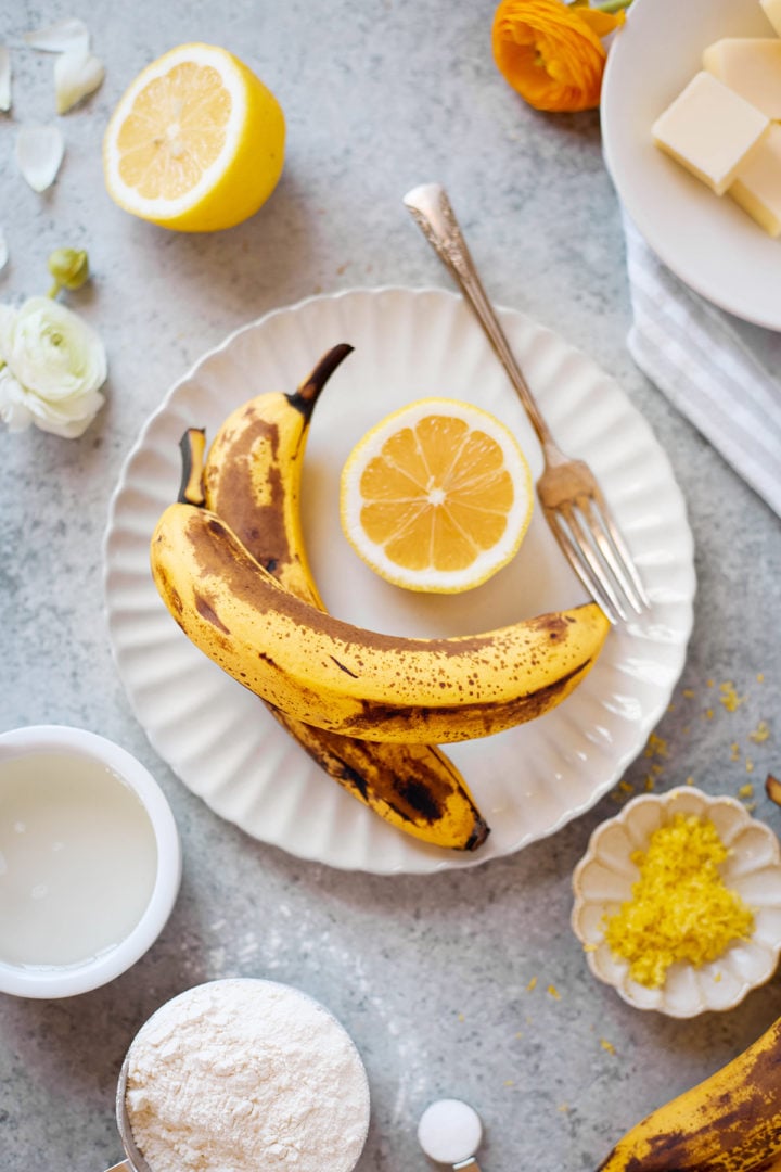Overhead view of ripe bananas and a halved lemon on a white plate, with a small fork resting nearby. Additional ingredients like flour, butter, lemon zest, and lemon juice are arranged around the scene—everything needed to make lemon banana bread from scratch.