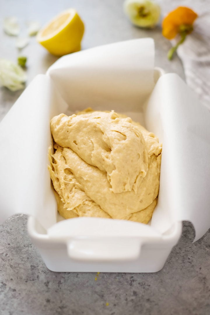 A close-up of thick banana bread batter with lemon poured into a parchment-lined loaf pan. A halved lemon and flowers are softly blurred in the background, evoking a light, spring-inspired feel.