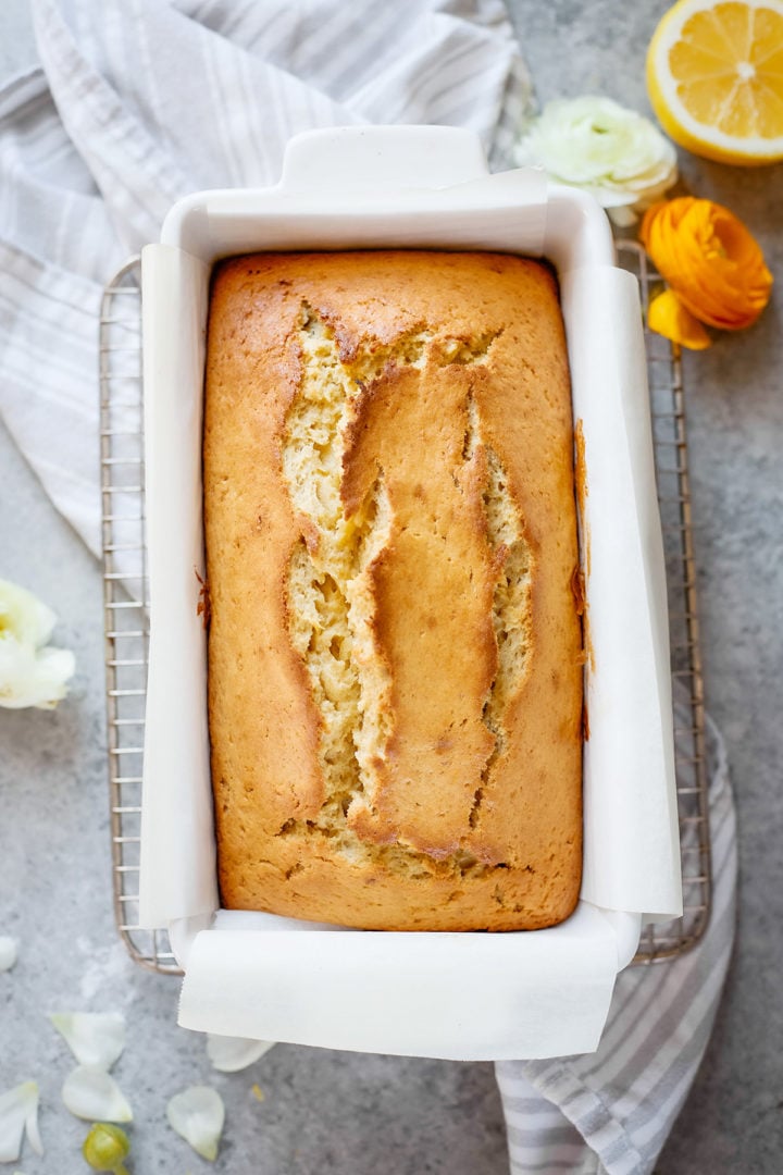 Freshly baked lemon banana loaf resting in a white ceramic pan lined with parchment. The top is perfectly cracked and golden brown. Lemon slices and spring flowers are arranged nearby for a bright, seasonal presentation.