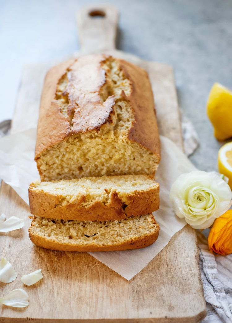 A close-up of a sliced lemon banana bread loaf showing its moist interior texture. Set on a wooden board with lemon halves and white and yellow flowers surrounding it—this homemade banana bread with lemon looks soft and fluffy.