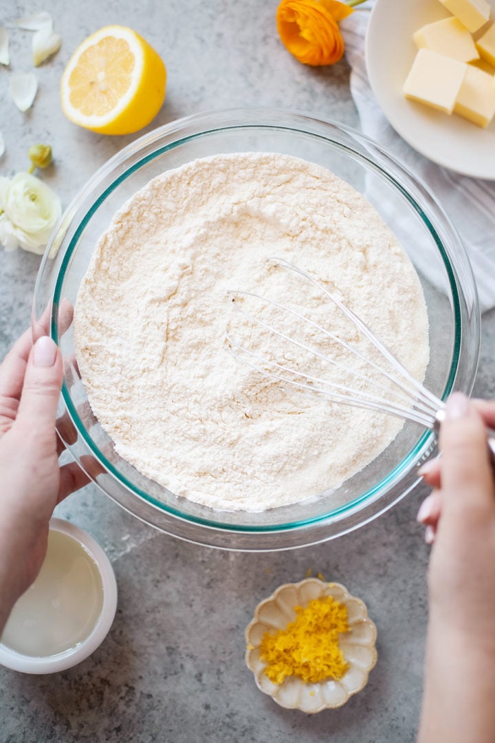 A glass bowl filled with flour being whisked by hand, surrounded by lemon zest, lemon juice, and softened butter. This step shows the dry ingredients being prepped to make lemon banana bread batter.